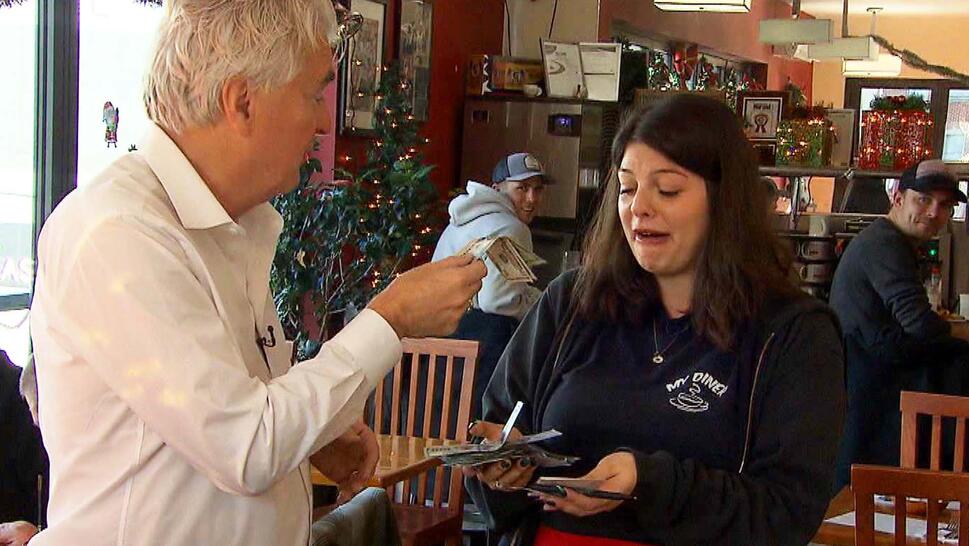 Man in a white button-up shirt handing cash to a waitress in a black long sleeved shirt that says 'My Diner' with a red apron around her waist.